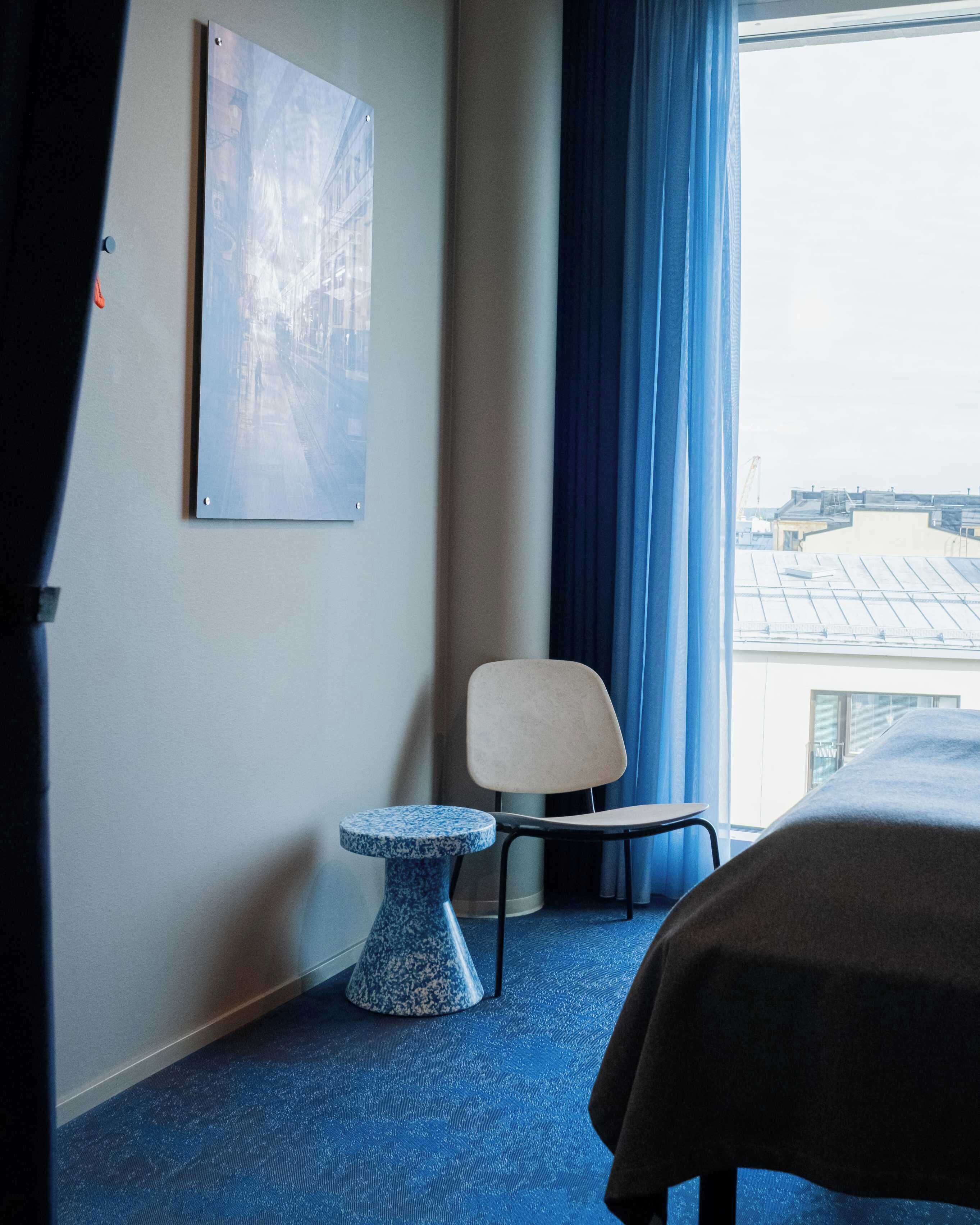 A hotel room with a bed, a chair, a small table, and a large window with a view of buildings.