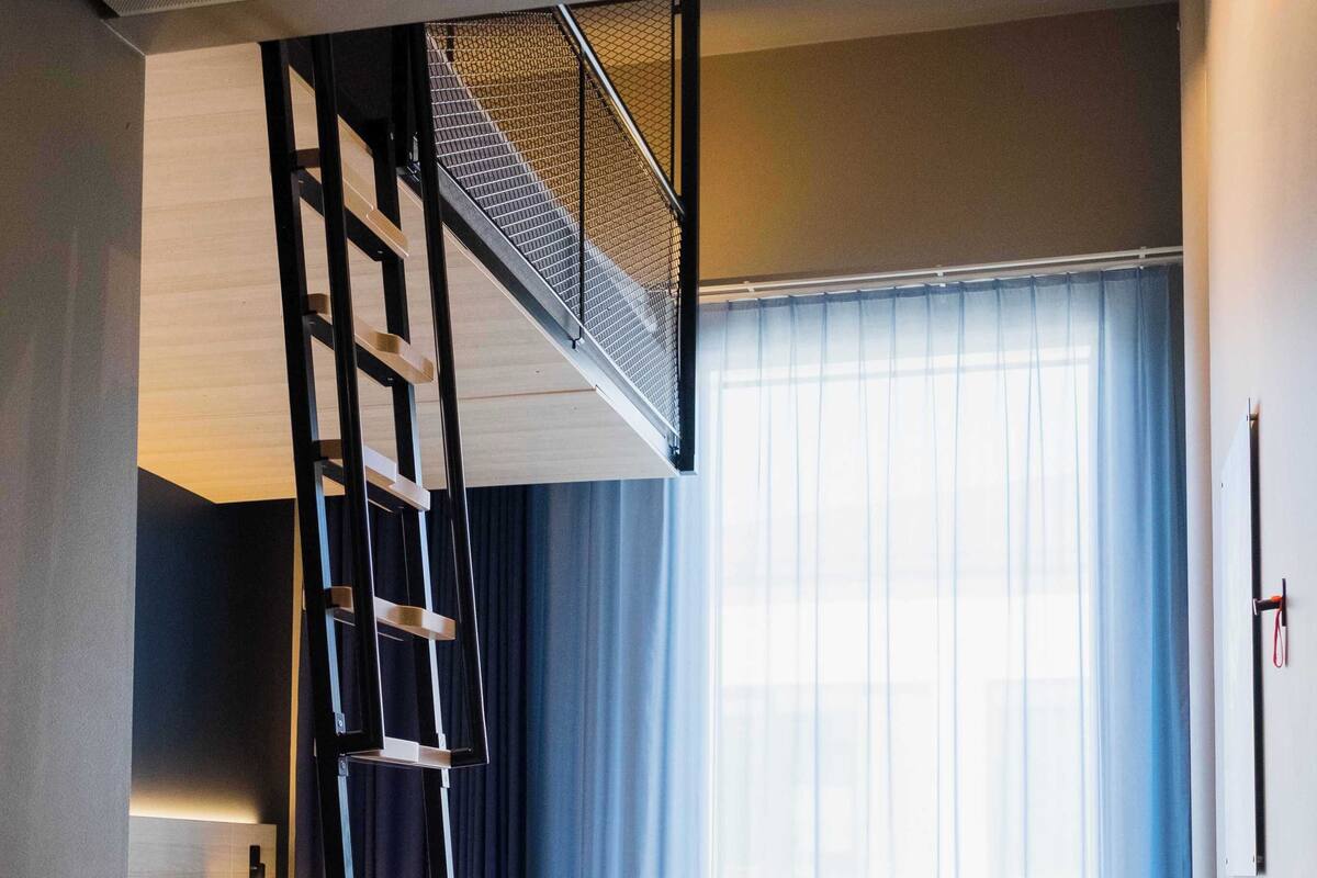 A modern hotel room with a bunk bed, a desk, a chair, and a large window with curtains.