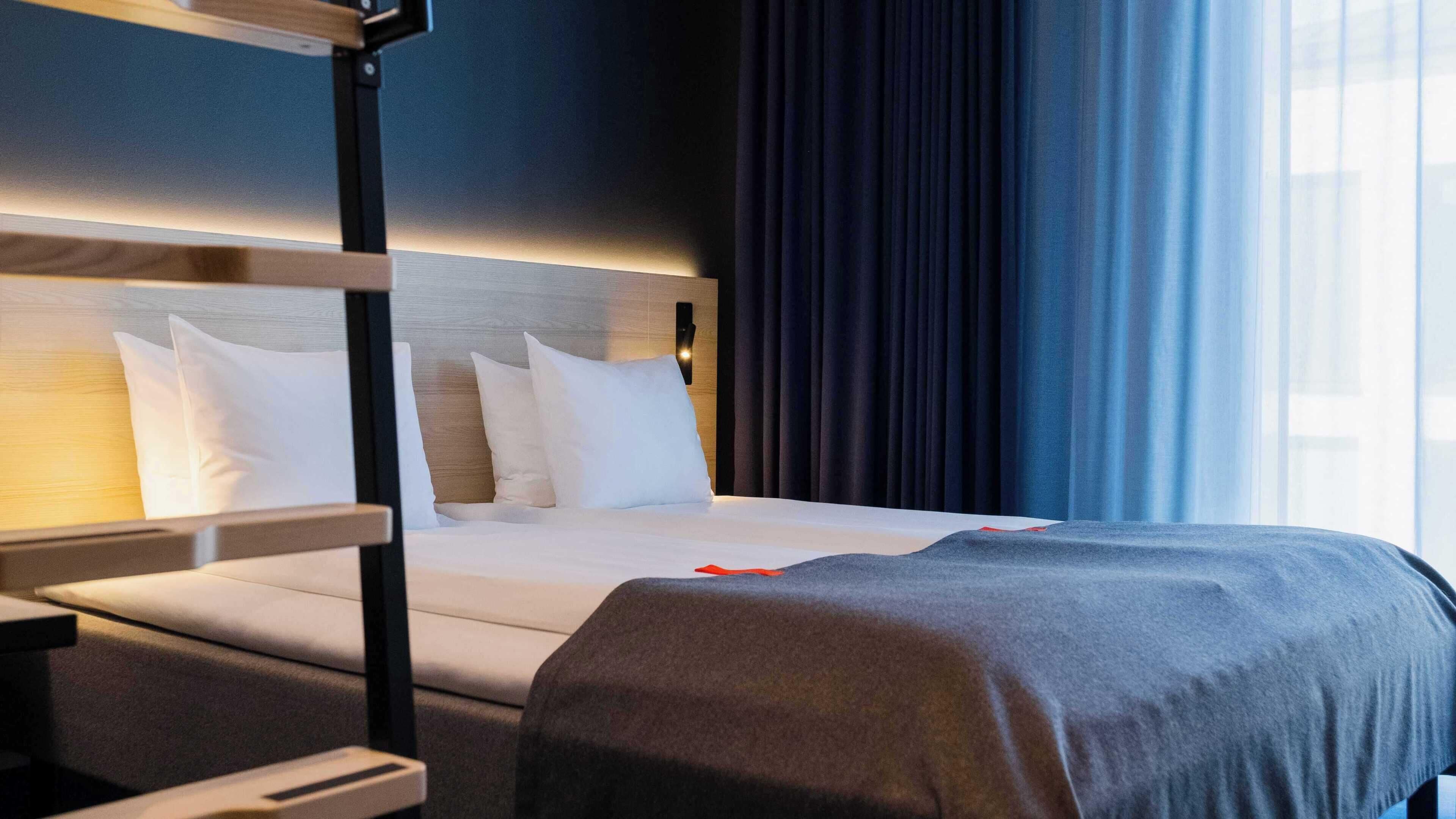 A hotel room with a bunk bed, a bedside table, a lamp, and a view of the outside through a window with curtains.