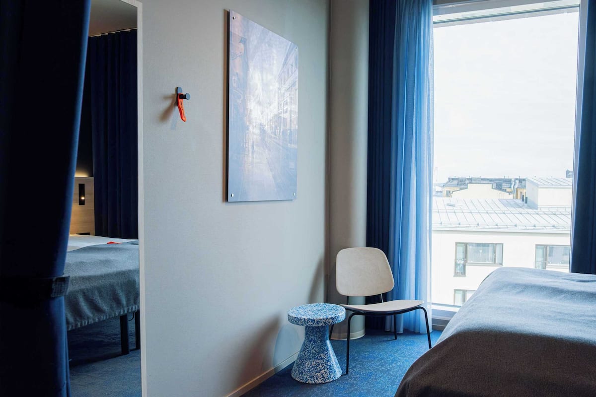 A hotel room with two beds, a small table, a chair, and a large window with a view of buildings.