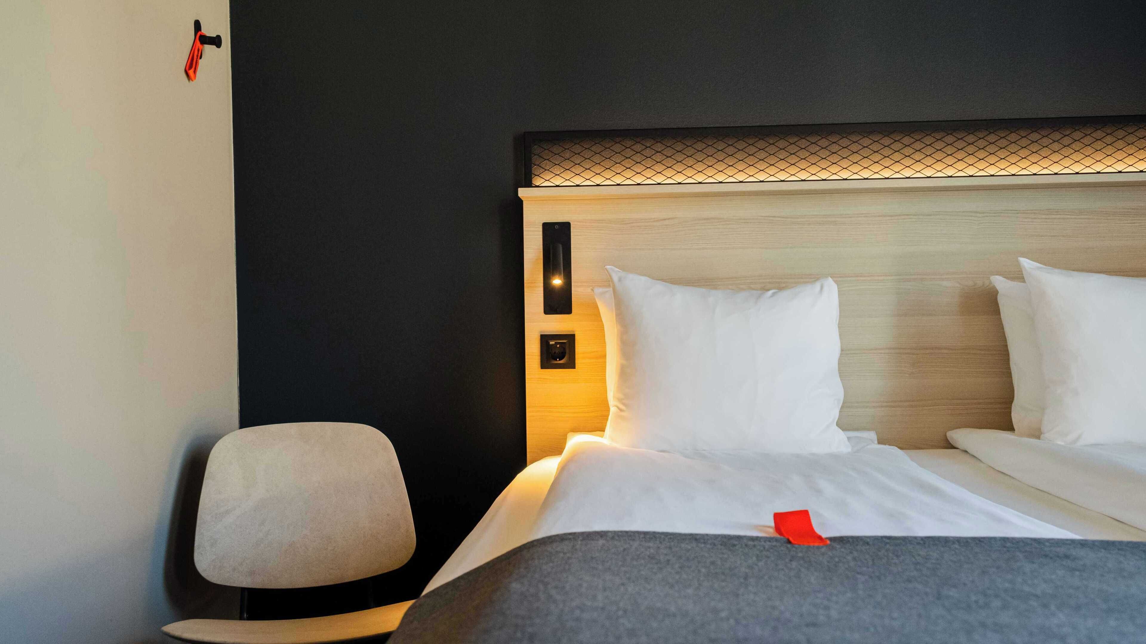 Una cama bien hecha con sábanas y almohadas blancas, una mesita de noche con una lámpara, y una silla.