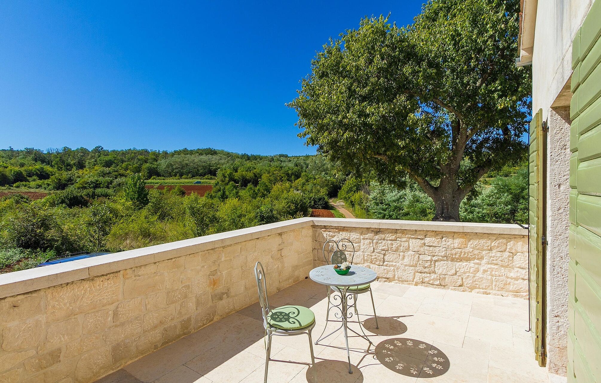 Un balcon avec un mur en pierre, une table, deux chaises et une vue sur un paysage verdoyant, avec des arbres et des collines.