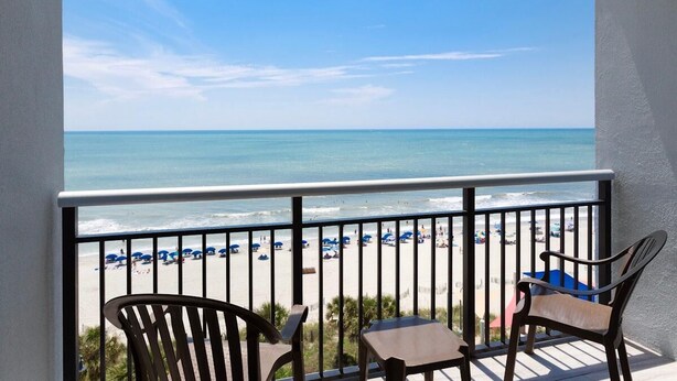 A balcony with a view of the beach, featuring two wooden chairs and a small table. - Beachside Serendipity Oceanfront 2 Bedroom (Myrtle Beach)