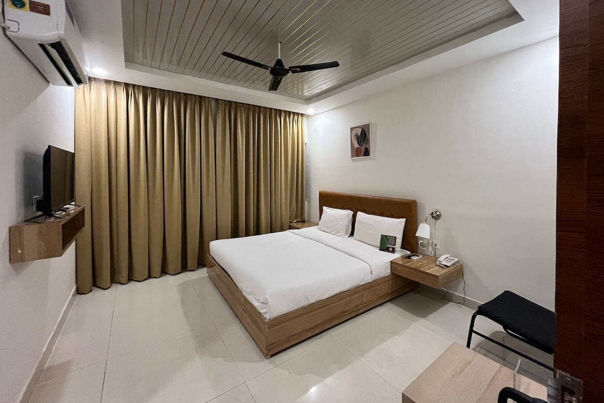 A hotel room with a bed, a ceiling fan, a TV, a bedside table with a phone, and a chair.