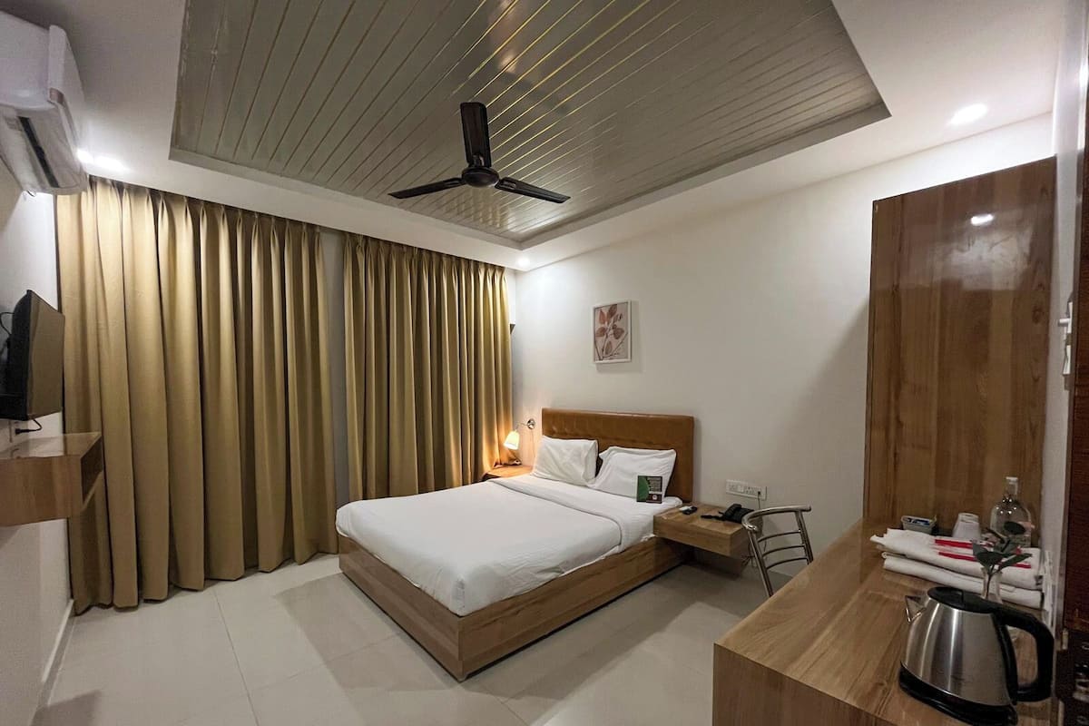 A hotel room with a bed, a desk with a kettle, a chair, a TV, and a ceiling fan.