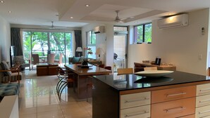 A modern kitchen with a central island, dining area, and a living space with a view of greenery. - BHA 1 Beachfront Haven - 2 bedroom (Mission Beach)