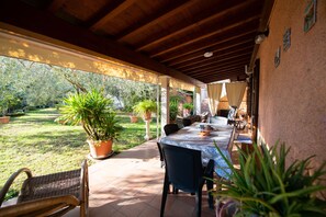 A covered patio with a table and chairs, surrounded by potted plants and a view of a garden.