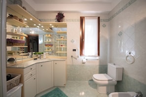 A modern bathroom with a toilet, bidet, and a sink area. There are shelves with toiletries and a mirror above the sink.