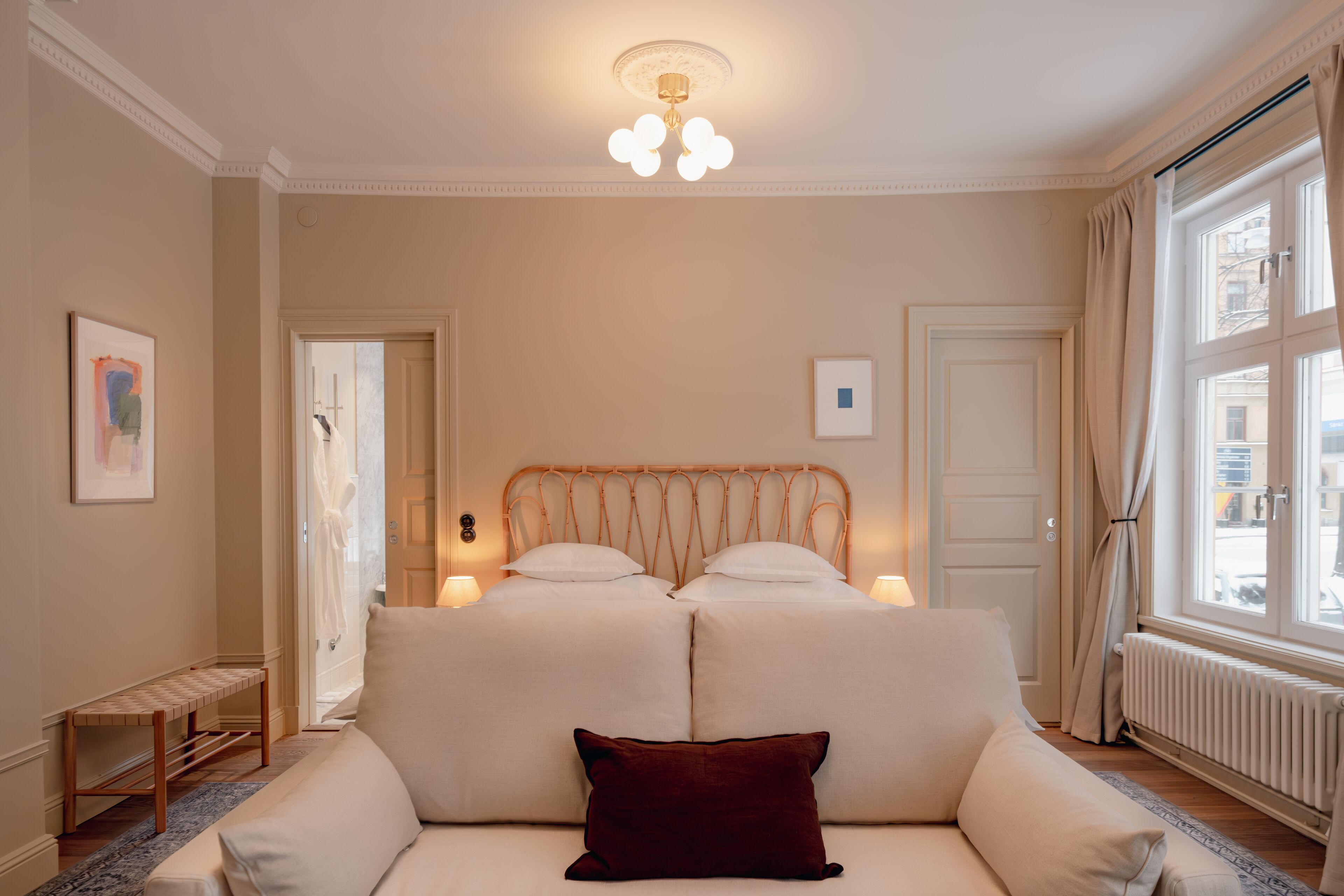 Una camera d'albergo con un letto, abat-jour, una finestra con tende, un termosifone e una piccola panca.