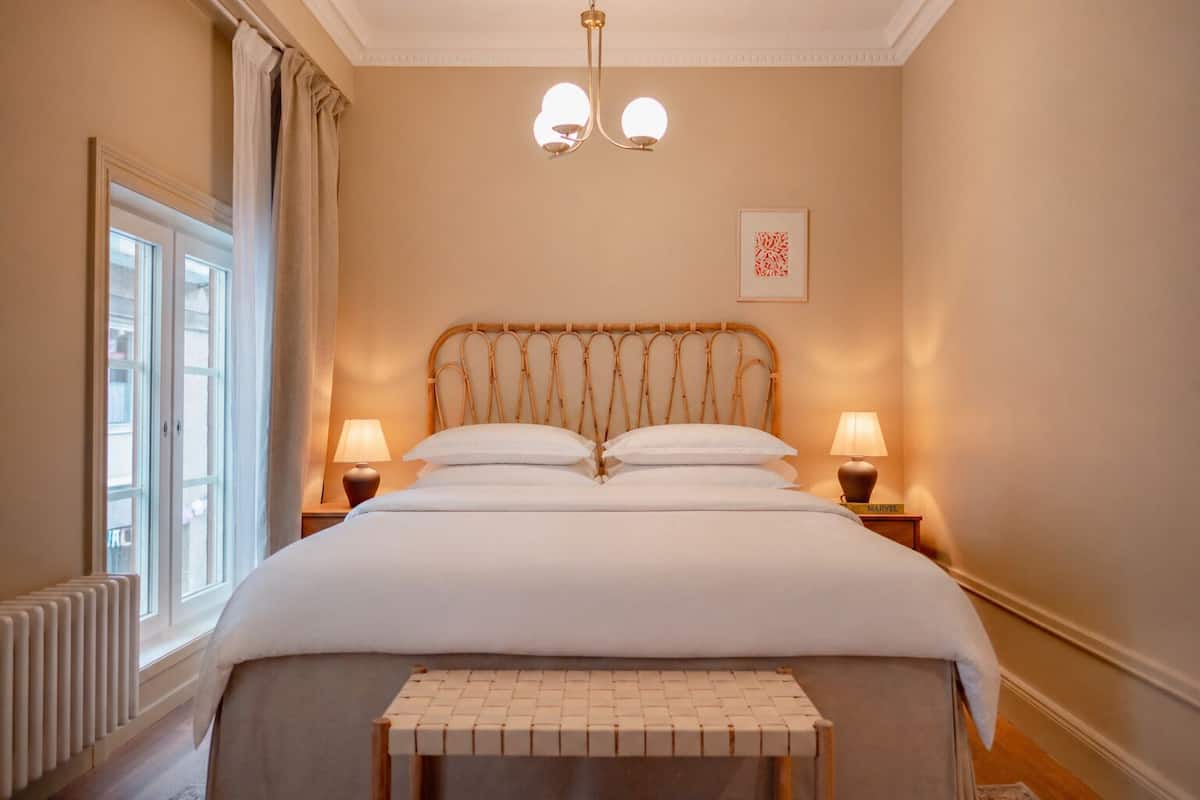 A neatly made bed with a headboard, two bedside lamps, a bedside table, and a window with curtains.