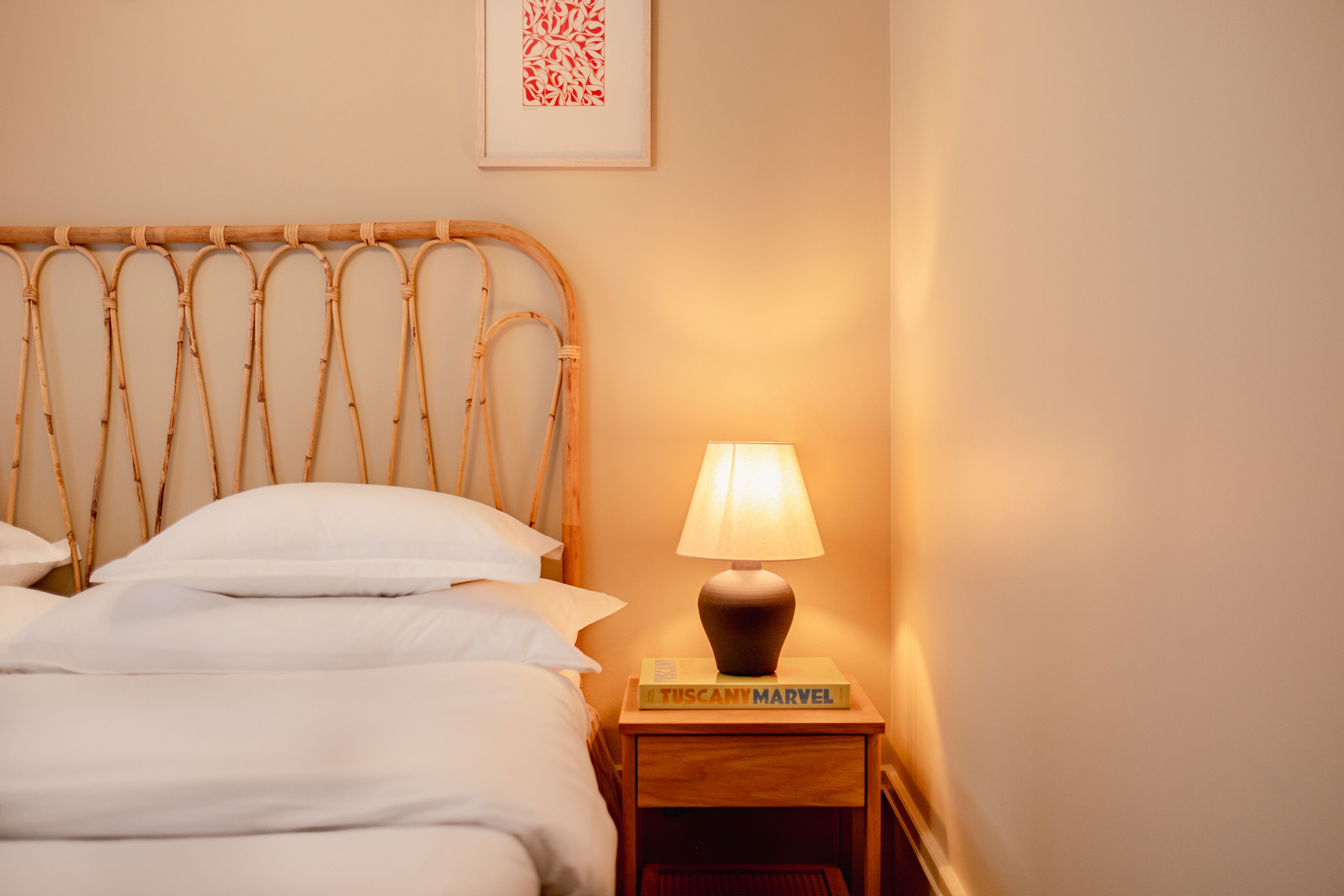 Een netjes opgemaakt bed met witte lakens, een houten hoofdbord, een bedlampje en een boek met de titel 'Tuscany Marvel' op het nachtkastje.