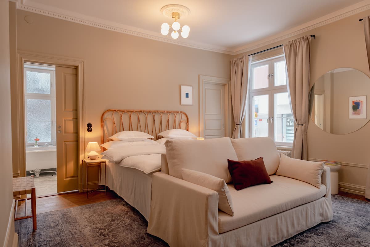 A bedroom with a large bed, a sofa, a bedside table, a lamp, and a window with curtains.