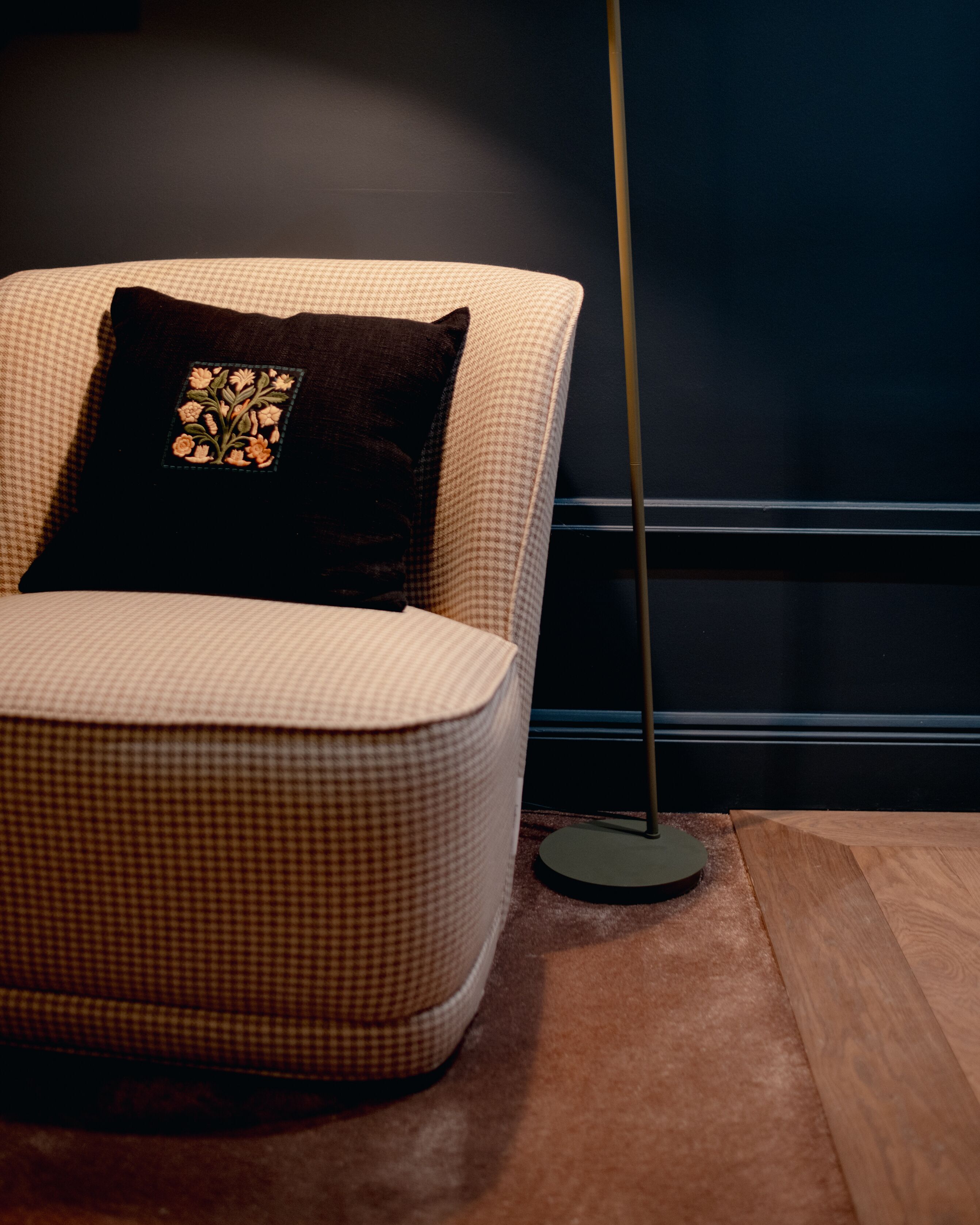 A checkered armchair with a floral pillow, a floor lamp, and a dark wall.