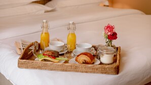 Una bandeja de desayuno ordenada sobre una cama con cabecera de cuadros, una botella de jugo de naranja, un croissant, un pequeño florero con flores y un periódico.