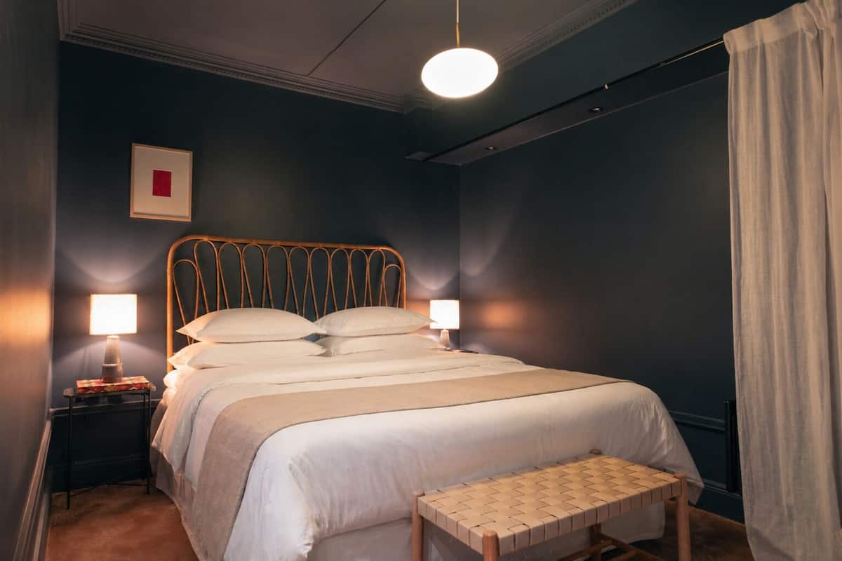 A bedroom with a bed, two bedside lamps, a bedside table, a small bench, and a single framed picture on the wall.