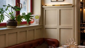 Un comedor con una mesa de madera dispuesta para cuatro personas, un banco con cojín rojo y una planta en maceta en el alféizar.
