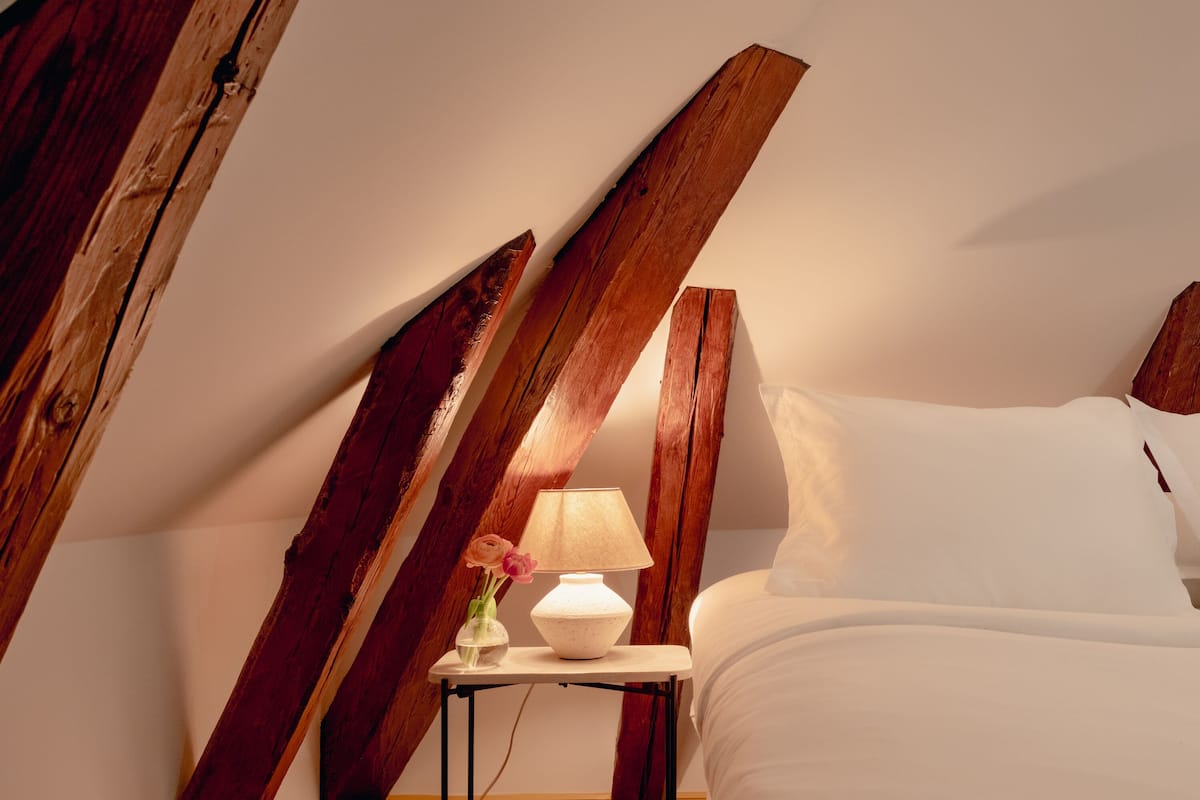 A bedroom with a sloped ceiling, wooden beams, a bedside table with a lamp, a vase with flowers, and a bed with white bedding.
