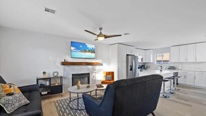 A modern living room with a fireplace, a ceiling fan, and a kitchen in the background. - Single Story 3BR w/ Pool, Hot Tub, Firepit & Grill (Henderson)