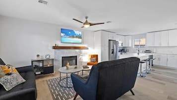 Una sala de estar moderna con chimenea, ventilador de techo y cocina al fondo.