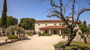 Un bâtiment en pierre, doté d’un toit en tuiles, entouré d’une allée gravillonnée et d’une végétation luxuriante.