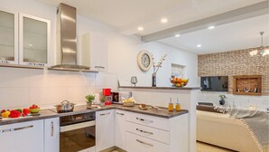 Une cuisine moderne avec des armoires blanches, une hotte de cuisinière en acier inoxydable et un four encastré. On y trouve un bar pour prendre le petit-déjeuner, avec un panier de fruits et une horloge au mur.