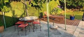 Relax in a serene outdoor space with a round table, four chairs, and a bench, surrounded by lush greenery and a metal fence.