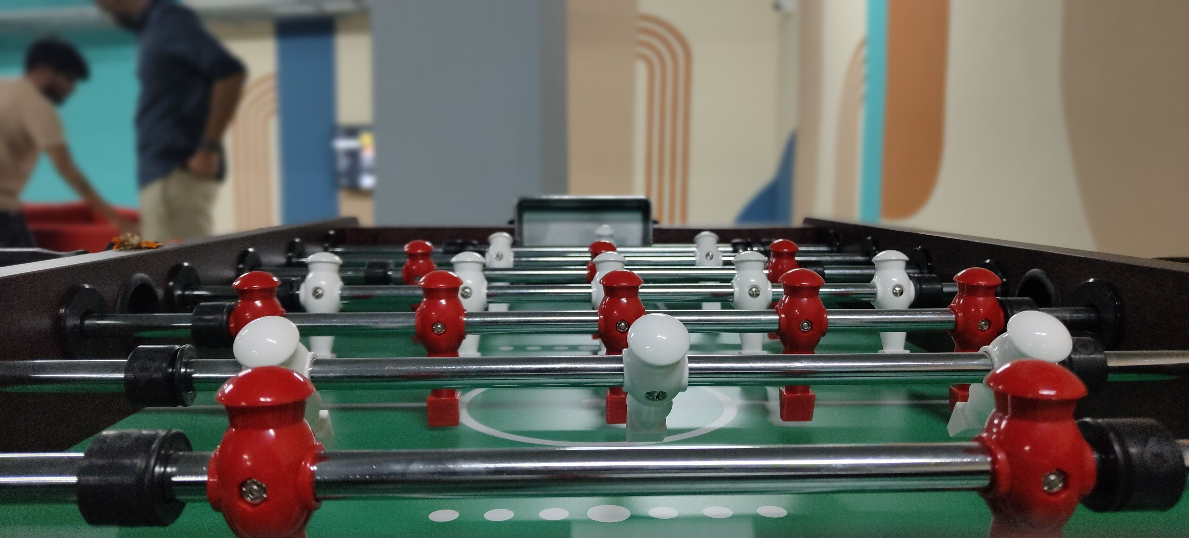 A foosball table with red and white figures, set in a room with a green playing surface and a visible score display.