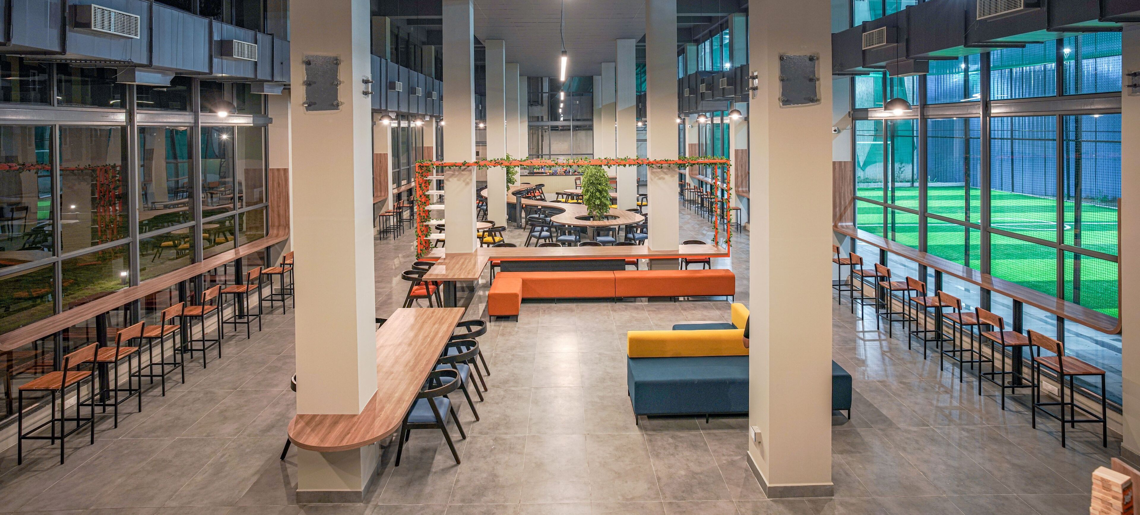 A spacious indoor area with multiple seating arrangements, including tables and benches, and large windows offering views of an outdoor sports field.