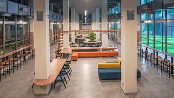A spacious indoor area with multiple seating arrangements, including tables and benches, and large windows offering views of an outdoor sports field.