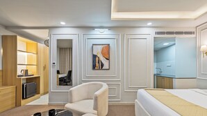 Una habitación de hotel con una cama, dos sillones, una mesita con dos tazas, un cuadro en la pared, un reloj y una zona de kitchenette.