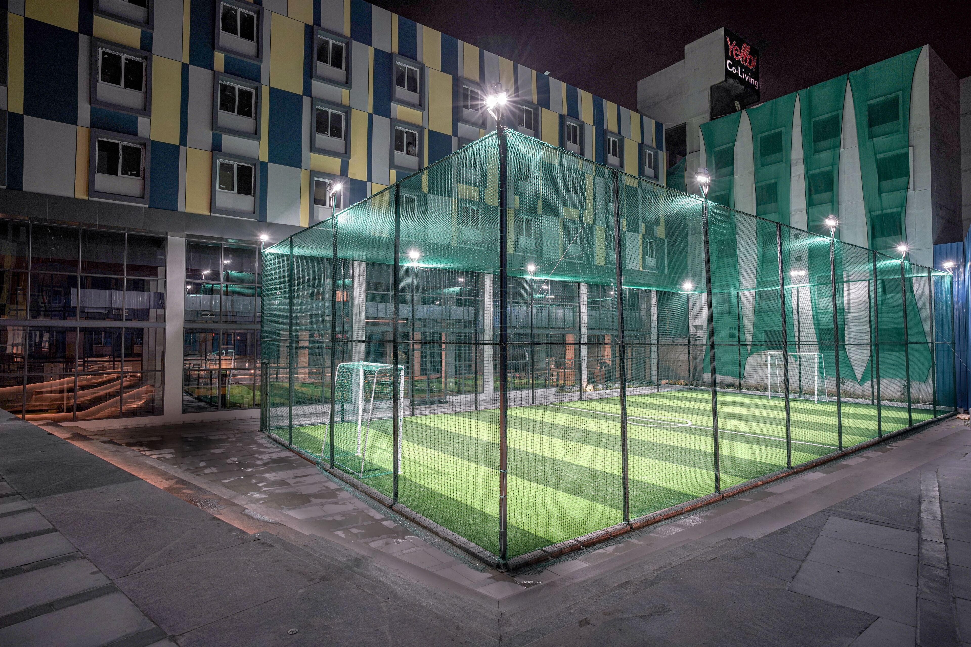 Eine Sportanlage mit einem umzäunten Bereich, einem Tor und einem Gebäude im Hintergrund mit Schachbrettmuster-Fassade.