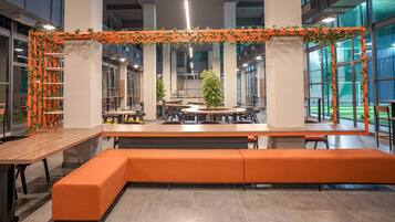 A modern indoor space with a central seating area, surrounded by tables and chairs, and large windows providing natural light.