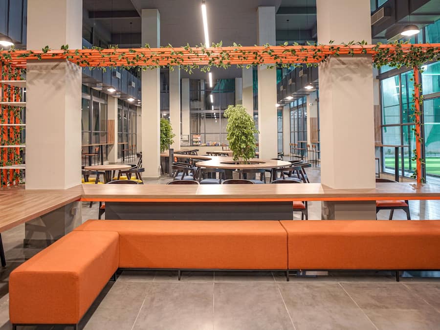 A modern indoor space with a central seating area, surrounded by tables and chairs, and large windows providing natural light.