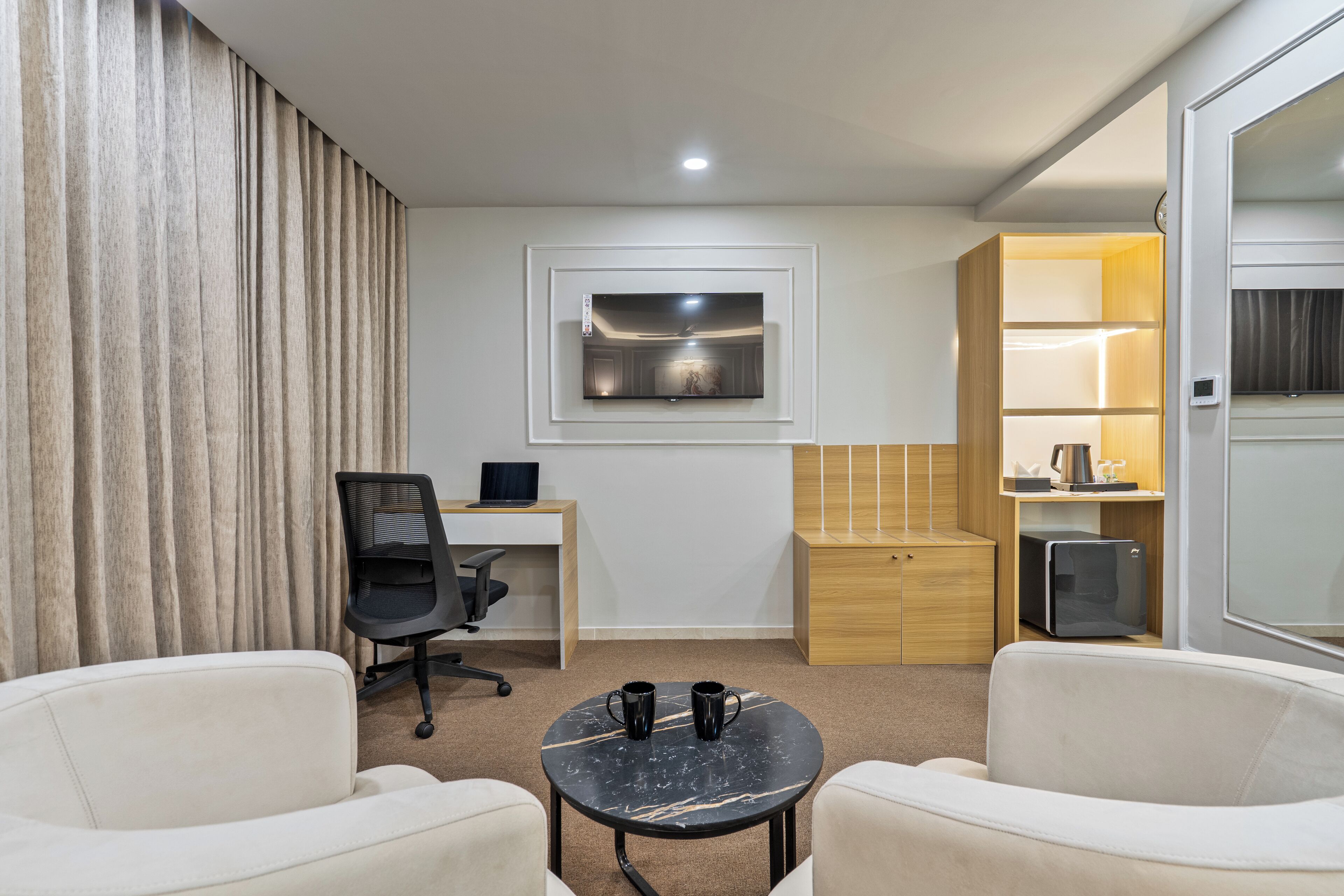 Una habitación de hotel moderna con escritorio, una silla de oficina, dos sillones, una mesita con dos tazas, un televisor montado en la pared y un minibar.
