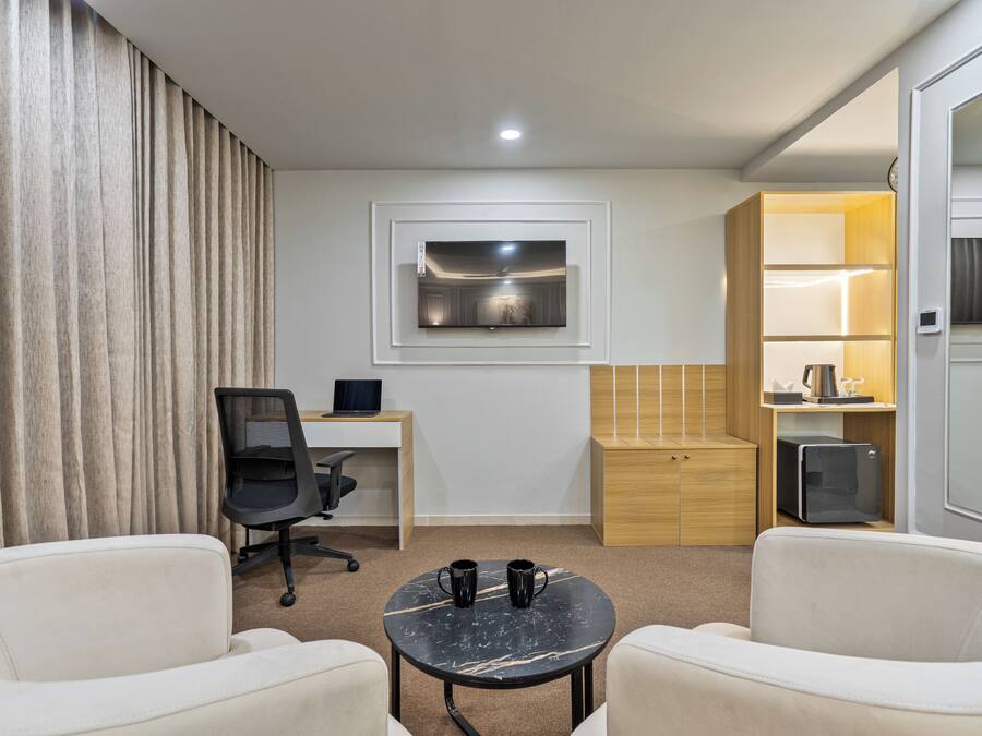 Una habitación de hotel moderna con escritorio, una silla de oficina, dos sillones, una mesita con dos tazas, un televisor montado en la pared y un minibar.