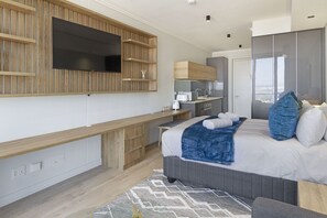 A modern bedroom with a large bed, a TV mounted on a wooden panel, a kitchenette, and a wardrobe with a mirror. - Ellipse Waterfall 31002 (Midrand)