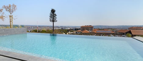 A rectangular swimming pool with clear blue water, surrounded by a tiled deck. In the background, there are residential buildings with brown roofs and a clear sky.