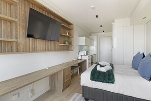 A modern hotel room with a wooden panel wall, a flat-screen TV, a kitchenette, a bed with blue pillows, and a green throw blanket. - Ellipse Waterfall 3902 (Midrand)