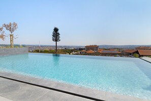 A rectangular swimming pool with clear blue water, surrounded by a tiled deck. In the background, there are residential buildings with brown roofs and a clear sky.