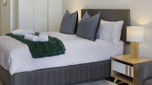Una cama bien hecha con sábanas blancas, un plaid verde y almohadas grises. Hay una mesita de noche con una lámpara y un cajón.