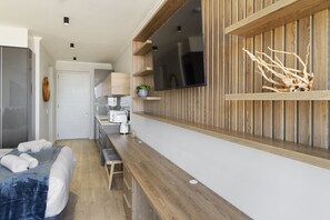 Une chambre d’hôtel moderne avec un mur d’accent en bois, un téléviseur à écran plat, un lit avec du linge de maison blanc, un petit coin cuisine avec une bouilloire et une plante sur le comptoir.