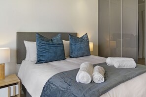 A neatly made bed with a grey headboard, two blue pillows, and two rolled white towels.