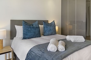 Een net opgemaakt bed met een grijs hoofdbord, twee blauwe kussens en twee opgerolde witte handdoeken.