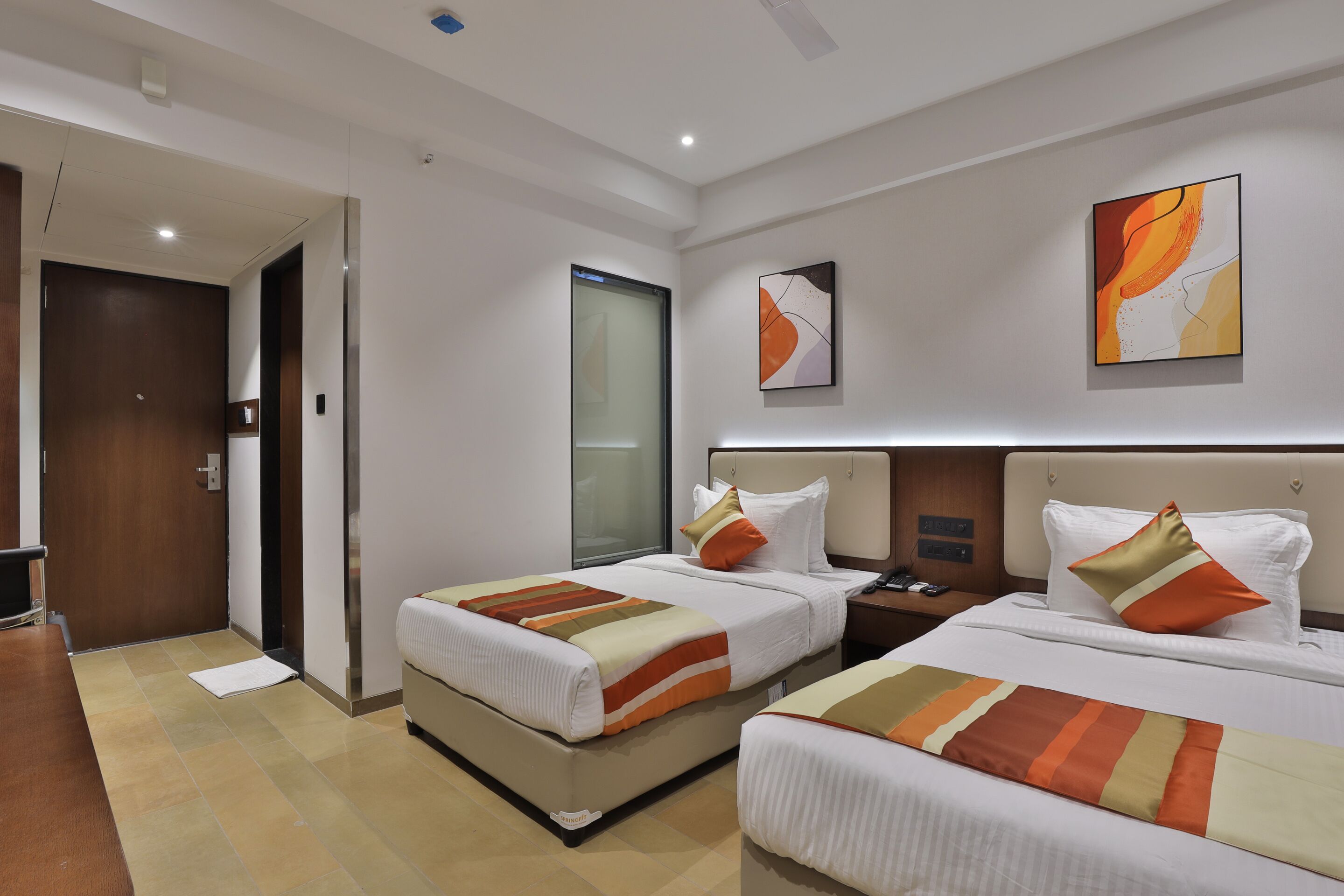 Habitación de hotel con dos camas, una puerta de madera, una mesita de noche y arte abstracto en las paredes.