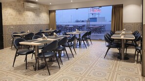 Restaurante com mesas e cadeiras dispostas para refeições, uma grande janela com vista e um aparelho de ar condicionado na parede.