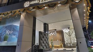 Entrada de hotel com design moderno, apresentando uma escadaria que leva ao saguão, um balcão de recepção e um grande painel digital na parede.