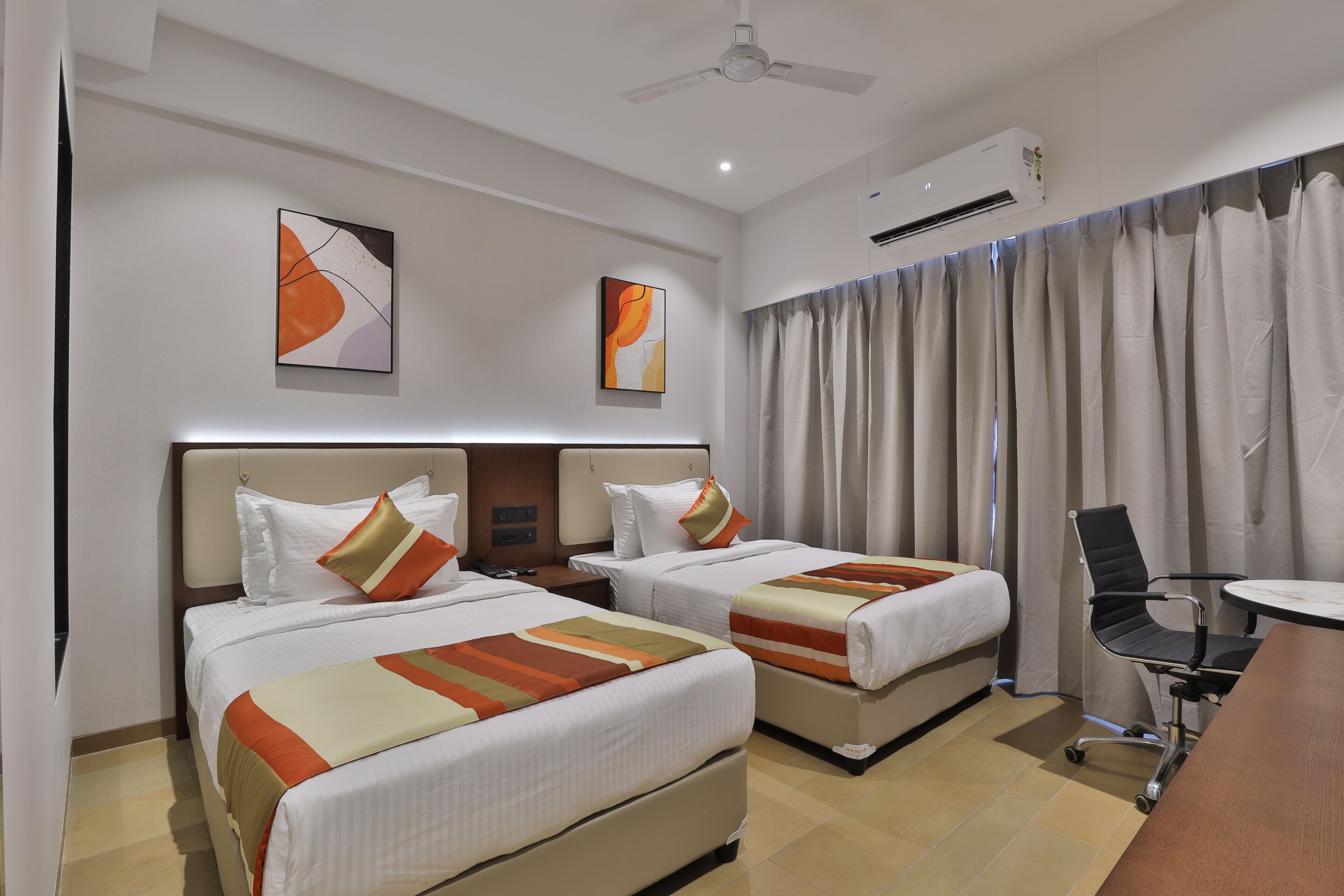 Habitación de hotel con dos camas, un escritorio, una silla, un equipo de aire acondicionado y arte abstracto en las paredes.