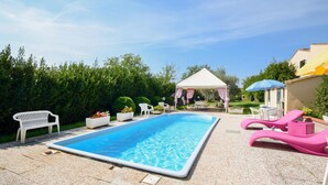 Une piscine rectangulaire bordée de blanc, entourée d’une terrasse pavée de petits galets. On y trouve deux chaises en plastique blanches et deux chaises longues roses. Une tente blanche, ornée de rideaux roses, est installée à l’arrière-plan.