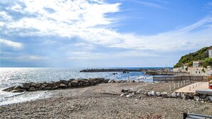 Une plage rocheuse avec une jetée en pierre qui s’avance dans la mer, un ciel dégagé et des bâtiments situés sur une colline.
