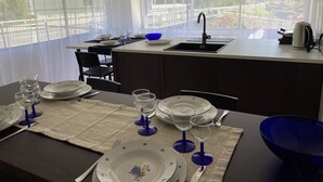 A dining area with a table set for four, a kitchen island with a sink, and a view of a road and greenery through large windows. - LA PLAGE large apartment with private beach (Pettenasco)
